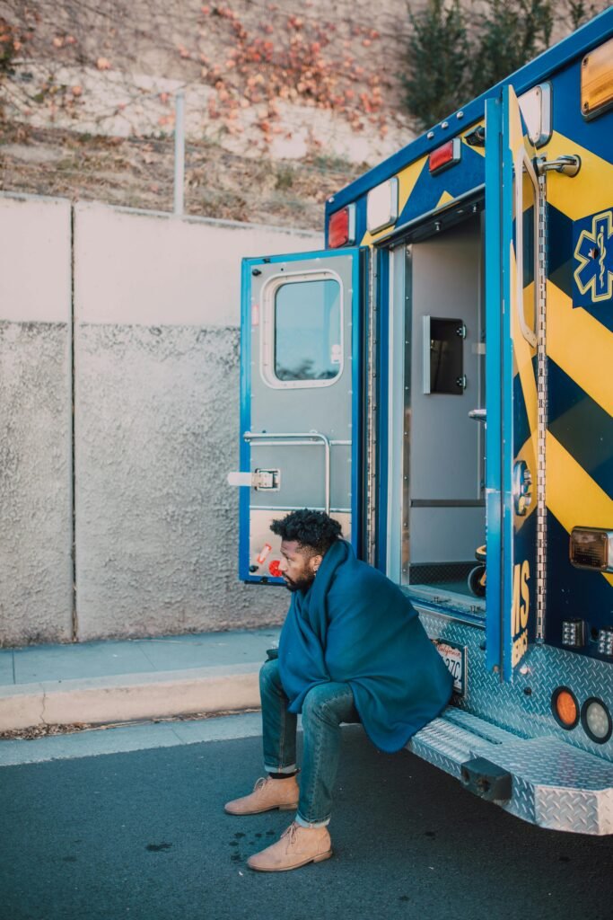 A man wrapped in a blanket sitting on the steps of an ambulance, appearing contemplative.