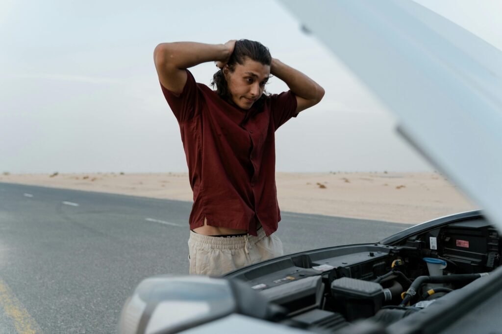 A frustrated man examining a broken car engine on a deserted road, showing stress and concern.
